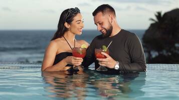 A couple in a swimming pool with drinks, overlooking the ocean.