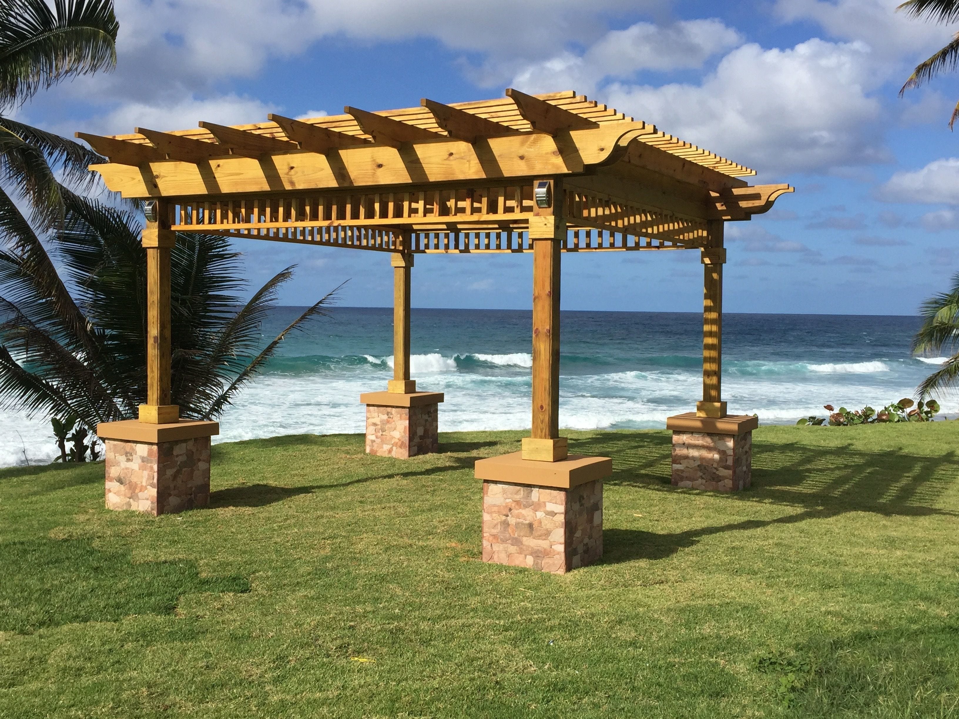 A wooden pergola with stone bases, overlooking a beach with waves and palm trees.
