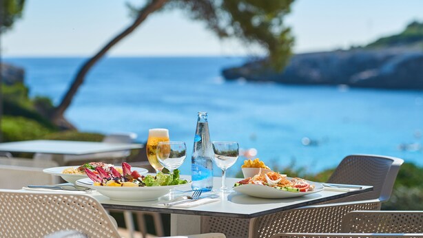 A table set with plates of food, glasses, and a water bottle, overlooking a coastal view.