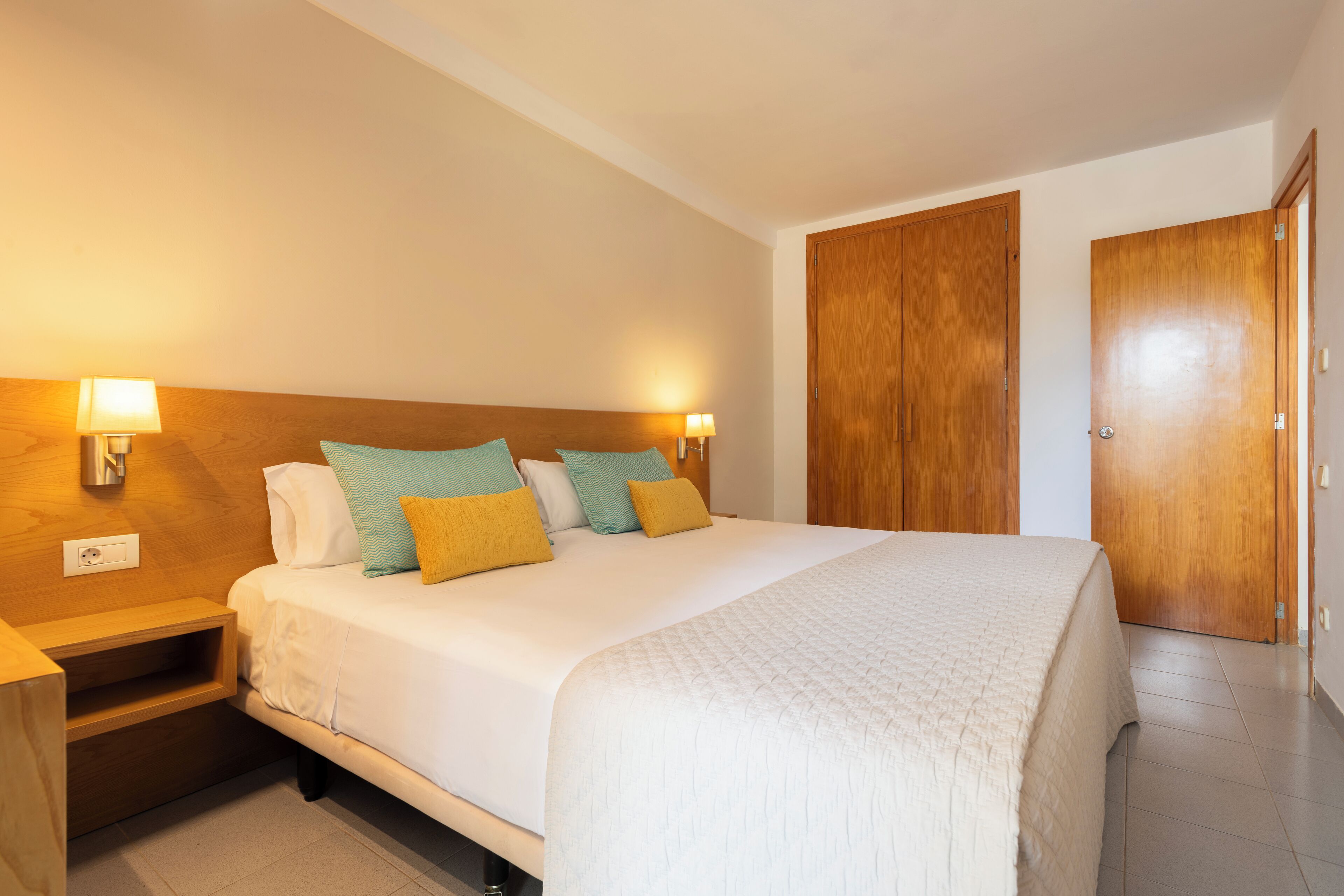Een keurig opgemaakt hotelkamer met een groot bed, houten hoofdeinde, twee bedlampen en een kledingkast.