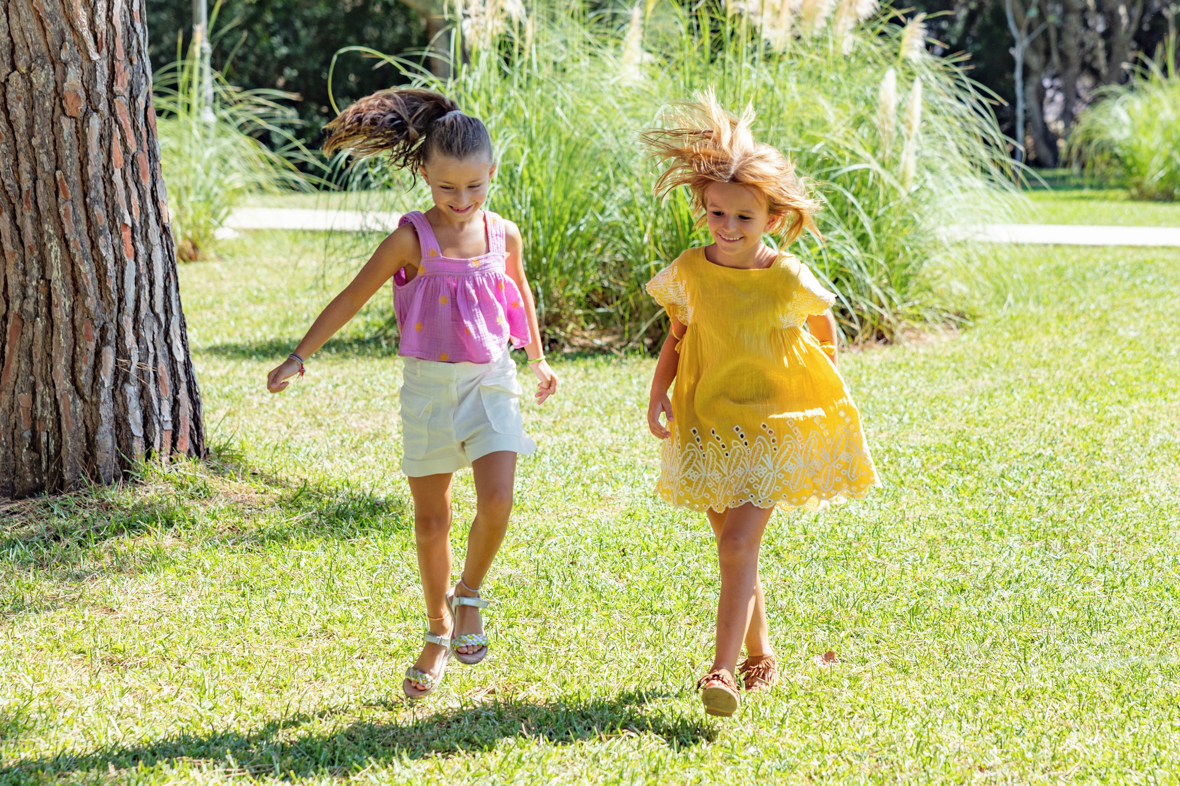 Duas crianças caminhando na grama, com uma árvore e um caminho ao fundo.
