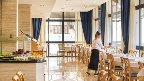 A spacious dining area with multiple tables and chairs, a buffet with fruits and beverages, and large windows with curtains.
