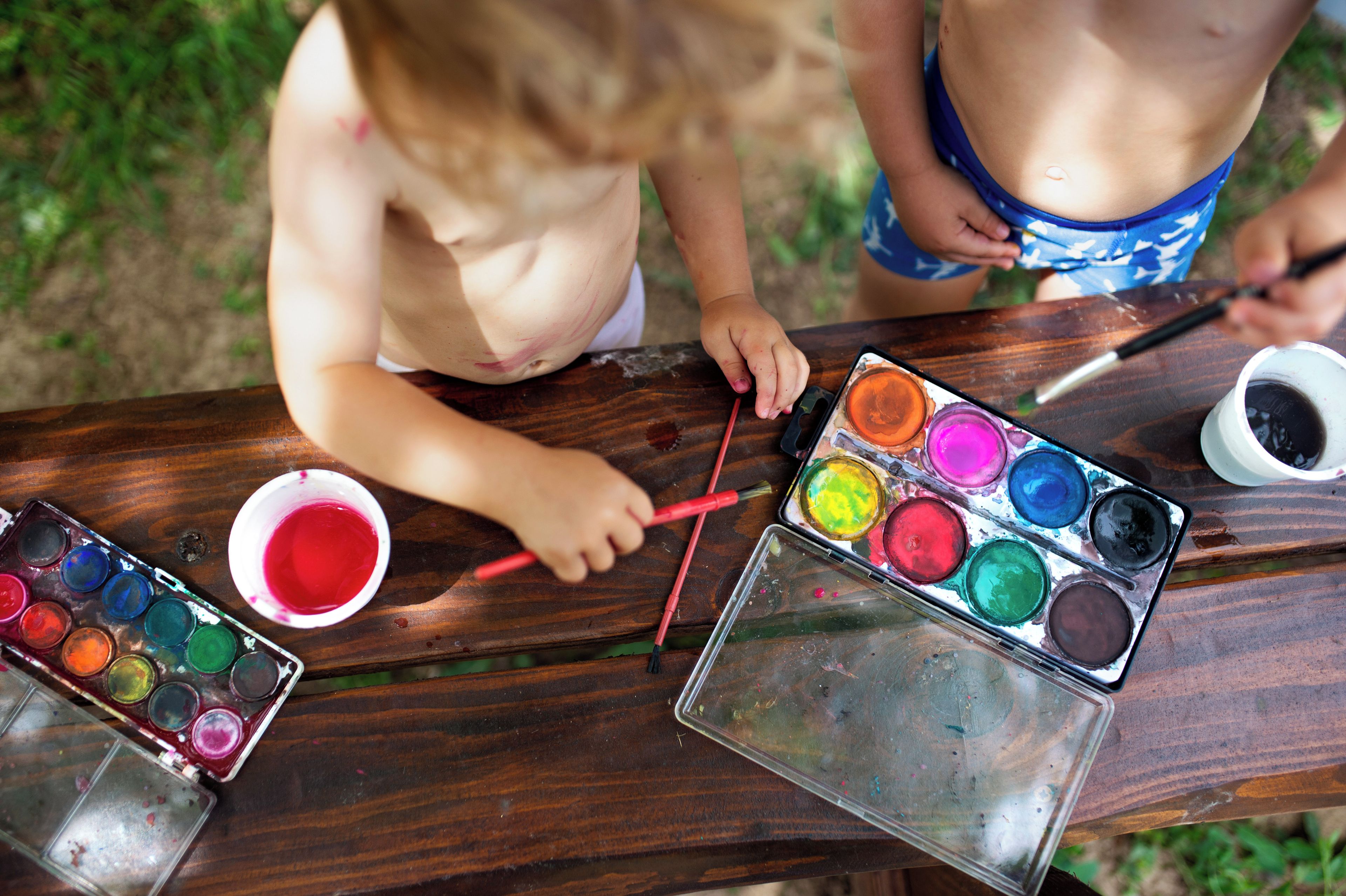 A wooden table with a child painting, a palette of colors, and a paintbrush.