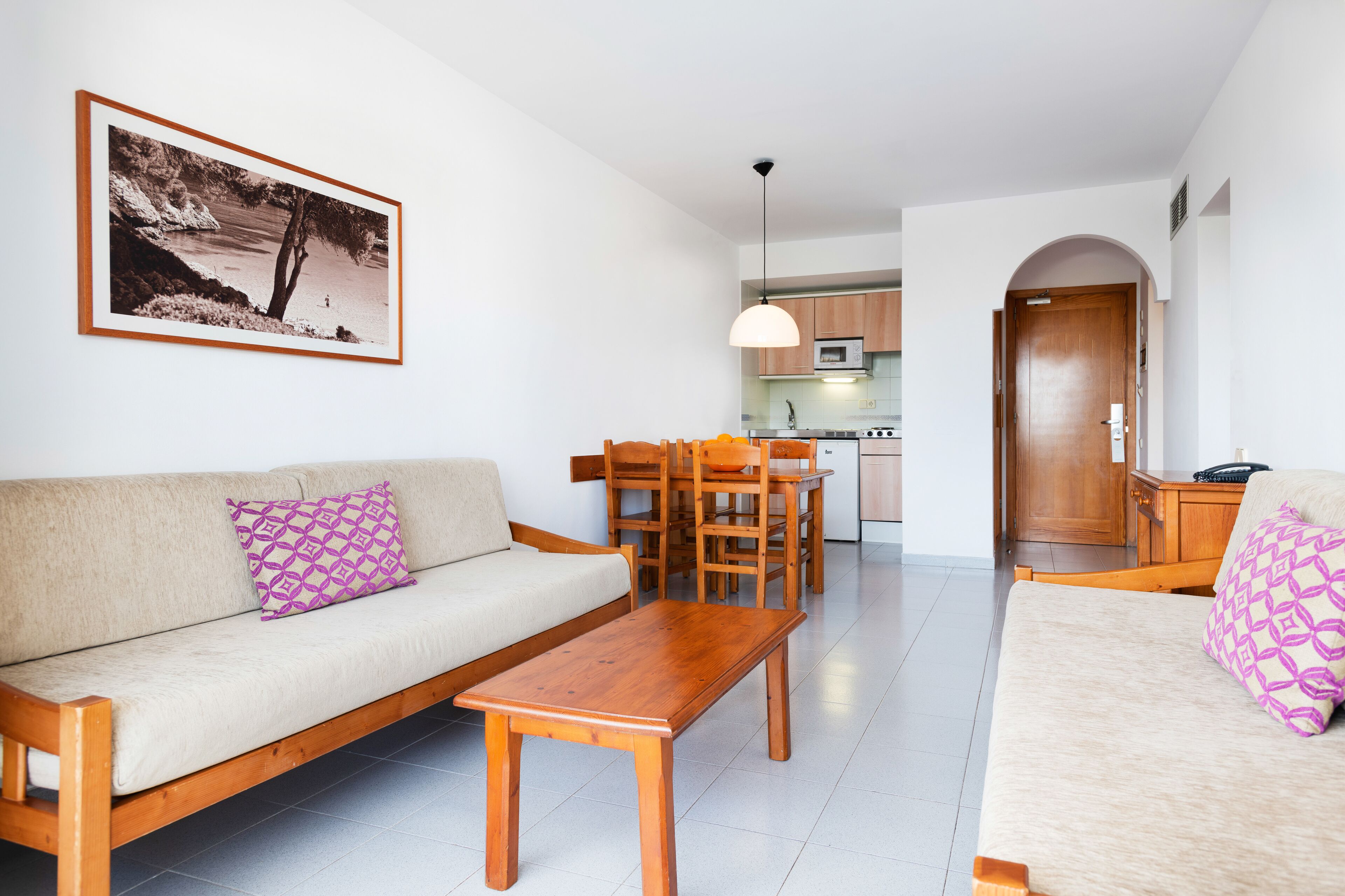 A living room with a sofa, a wooden coffee table, and a dining table with chairs. There is a kitchen in the background with cabinets and a microwave.