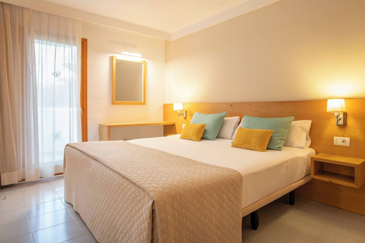 A hotel room with a large bed, two bedside tables, a nightstand, a mirror, and a window with curtains.