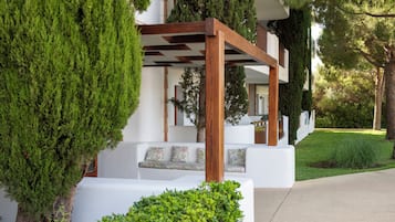 Un edificio moderno con fachada blanca, pérgola de madera y un sofá blanco con cojines estampados.