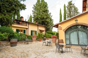 Un patio con suelo de piedra, mobiliario exterior, plantas en macetas y un edificio con balcón.