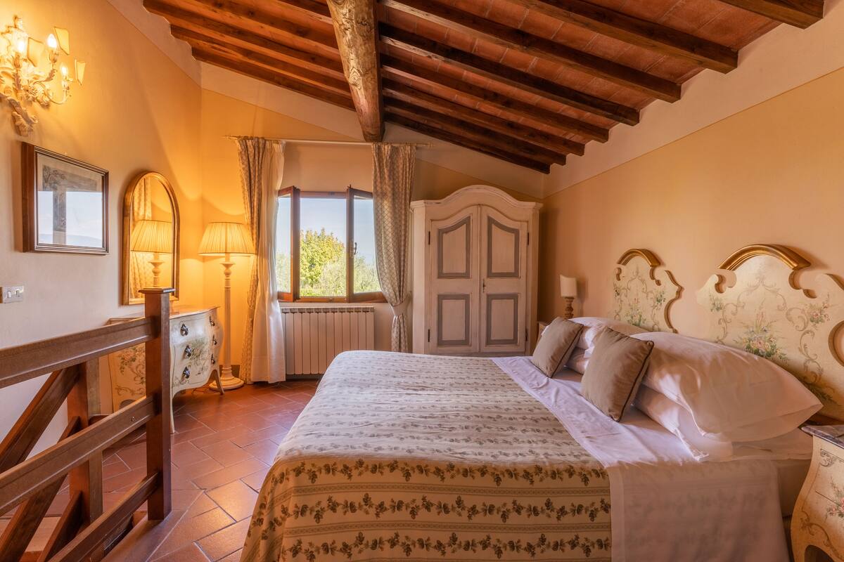 Une chambre à coucher avec un grand lit, des poutres apparentes en bois au plafond, une commode et une fenêtre avec des rideaux.