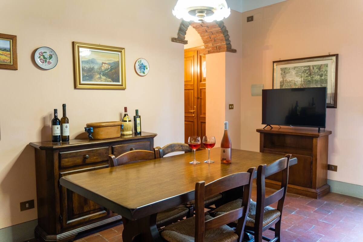 Un coin repas avec une table en bois, des chaises et un téléviseur posé sur un meuble. On peut voir des bouteilles de vin et des verres sur la table.