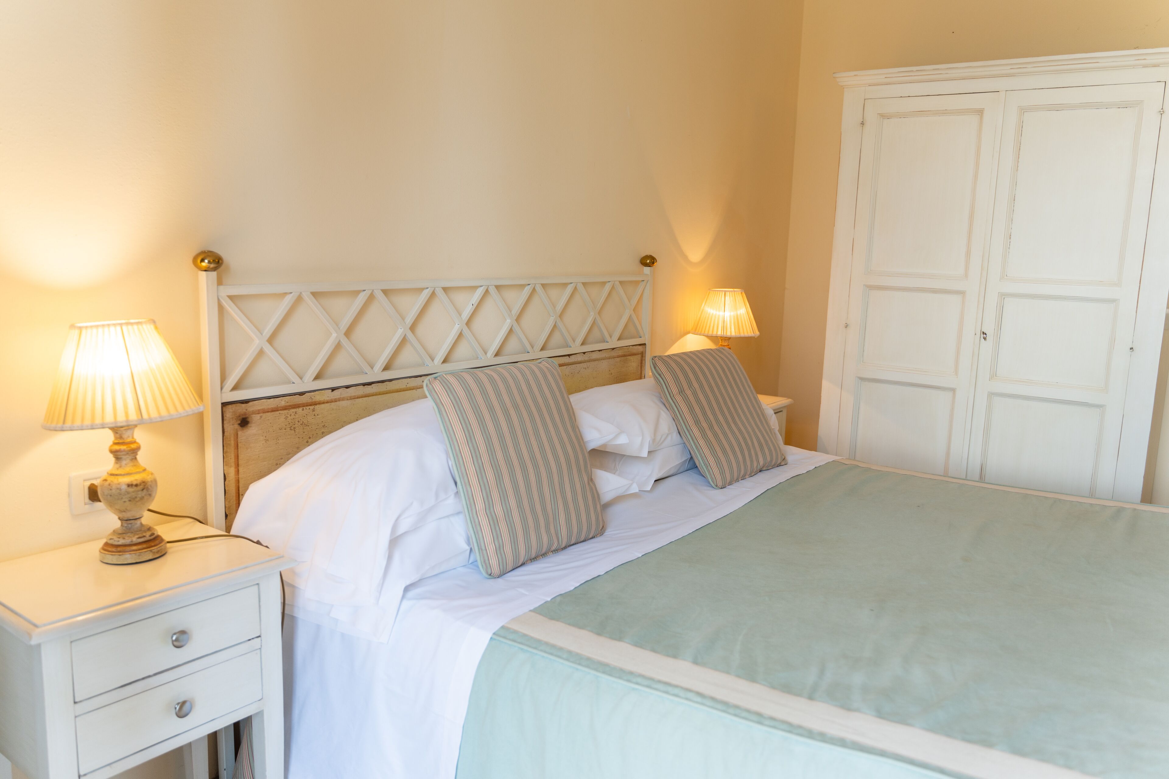 Una cama bien hecha con almohadas, una lámpara de noche y un armario blanco en una habitación con paredes lisas.