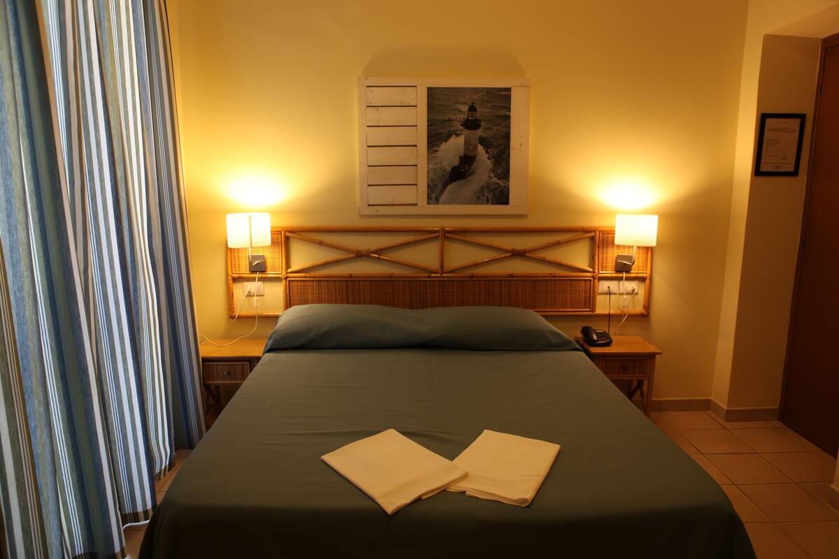 A hotel room with a bed, two bedside lamps, a wall-mounted picture, and a window with curtains.