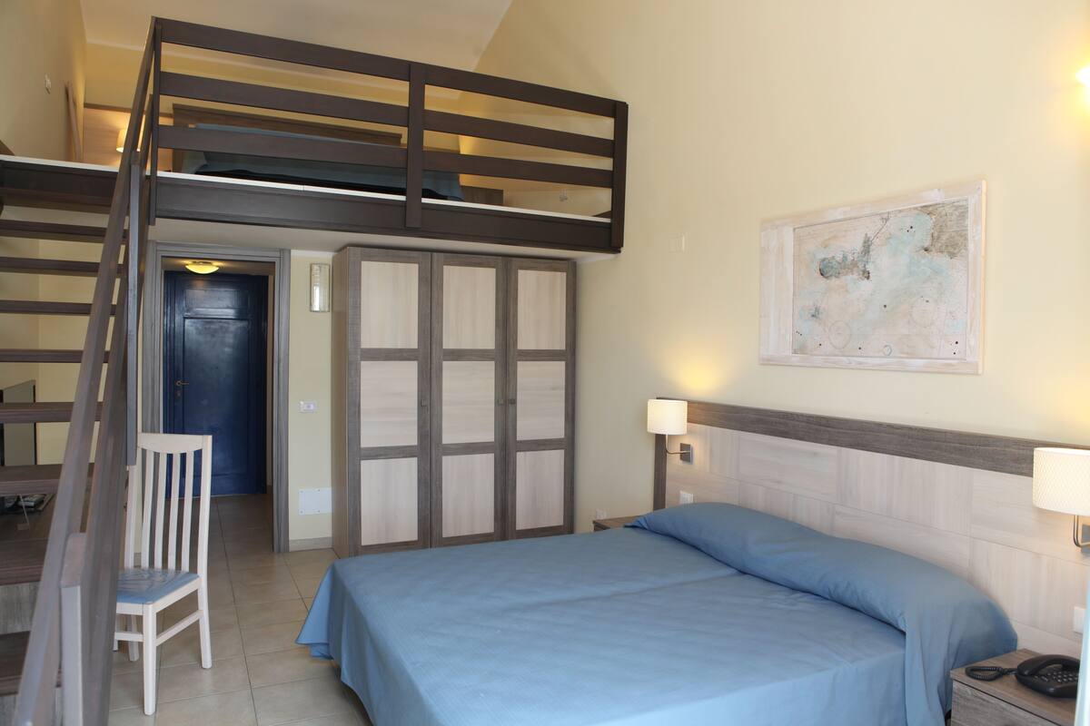 A bedroom with a bunk bed, a single bed, a chair, a bedside table with a lamp, and a wall-mounted artwork.