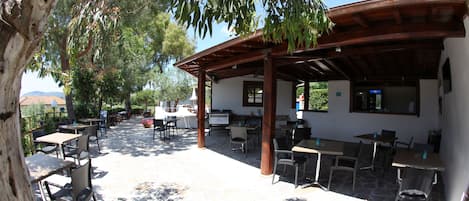 Un patio al aire libre con piso de piedra, mesas y sillas de madera, y una zona de estar techada.