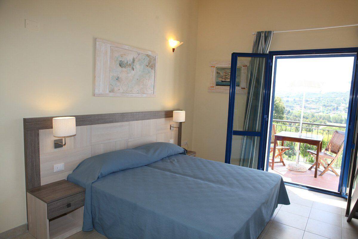 A bedroom with a bed, bedside lamps, a nightstand, a window with a view, and a balcony with chairs.