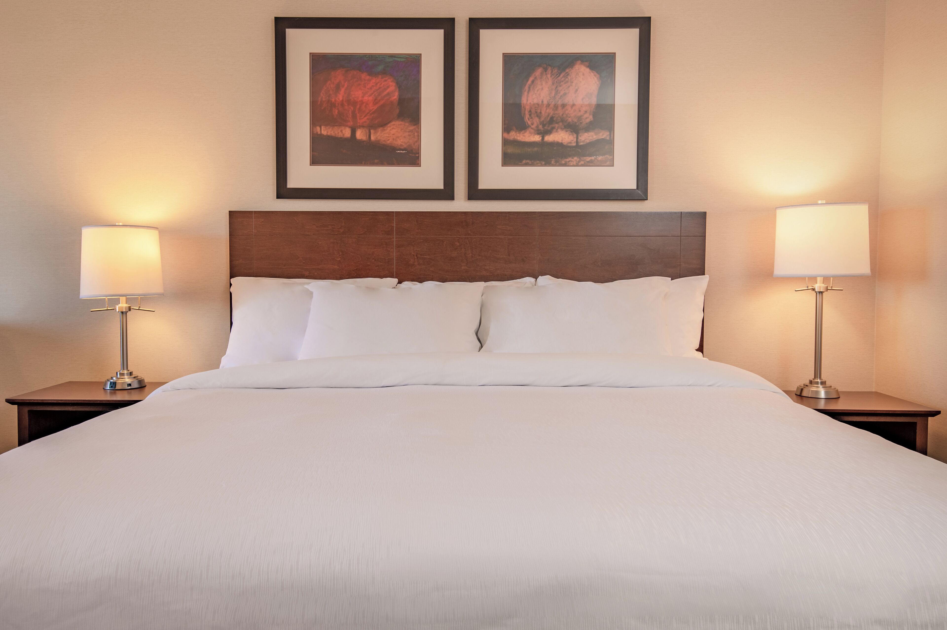 A neatly made bed with white linens, two framed pictures on the wall above, and two bedside lamps on either side.