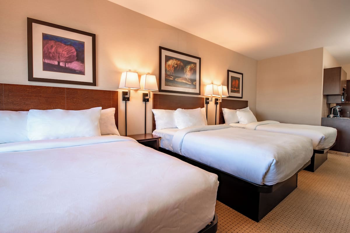 Habitación de hotel con tres camas, cada una con ropa blanca y cabeceros. Hay lámparas de pared y cuadros enmarcados sobre las camas.
