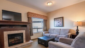A living room with a fireplace, a flat-screen TV mounted above it, a gray sofa, a coffee table, and a lamp. - Canalta Hotel High River (High River)