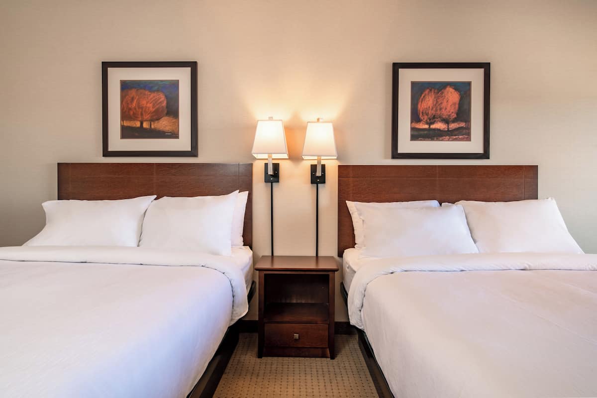 Habitación de hotel con dos camas, cada una con ropa de cama blanca, cabecero de madera y mesita de noche con lámpara.