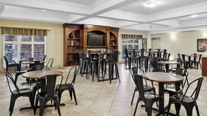 A spacious dining area with multiple tables and chairs, a wooden entertainment unit, and large windows with curtains. - Canalta Hotel High River (High River)