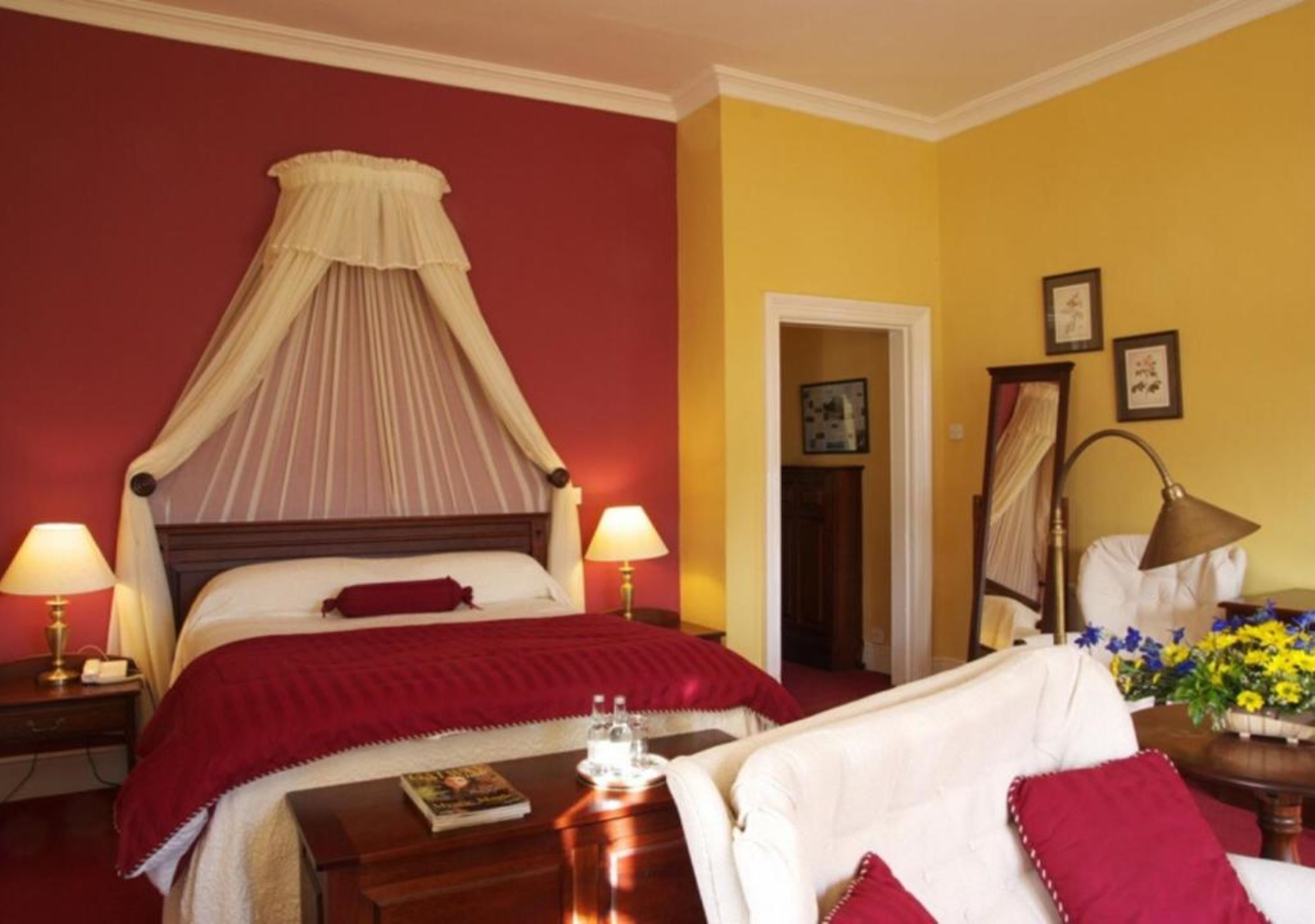 A hotel room with a four-poster bed, two bedside lamps, a bedside table with a book and glasses, a chair with a red cushion, and a vase with flowers.