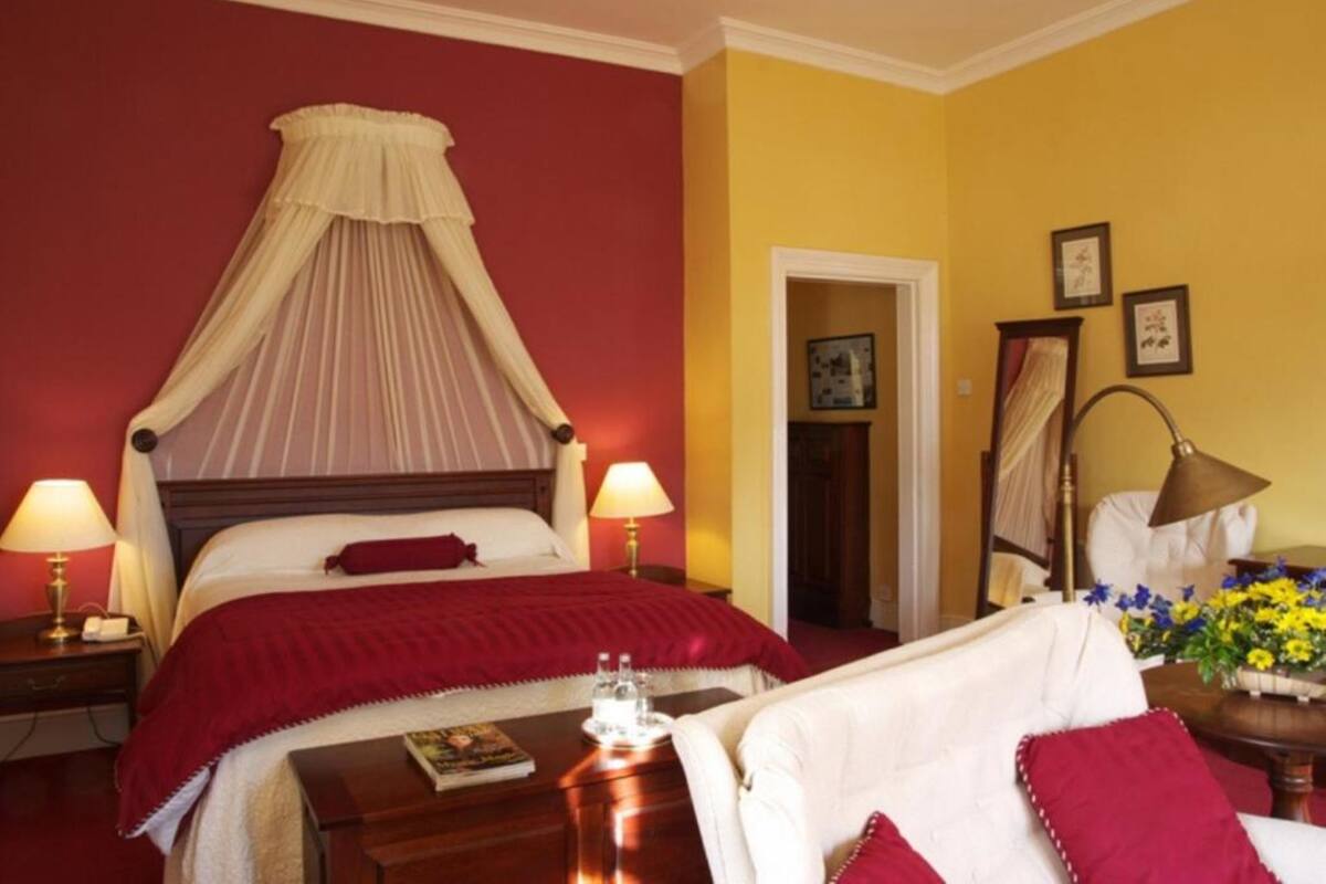 A hotel room with a four-poster bed, two bedside lamps, a bedside table with a book and glasses, and a floral arrangement on the right.
