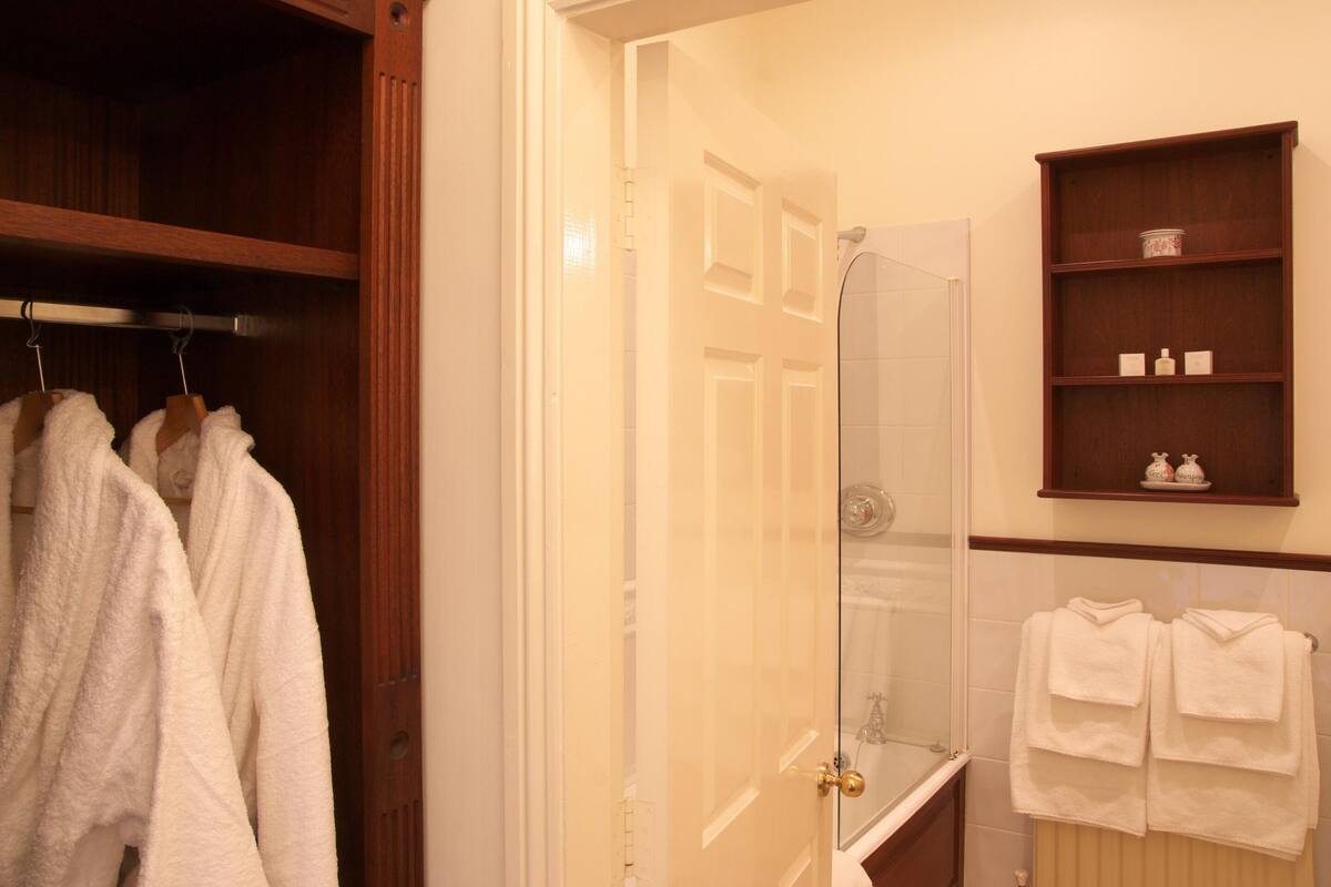 A bathroom with a glass shower door, a wooden shelf with toiletries, and two towels hanging on the wall.