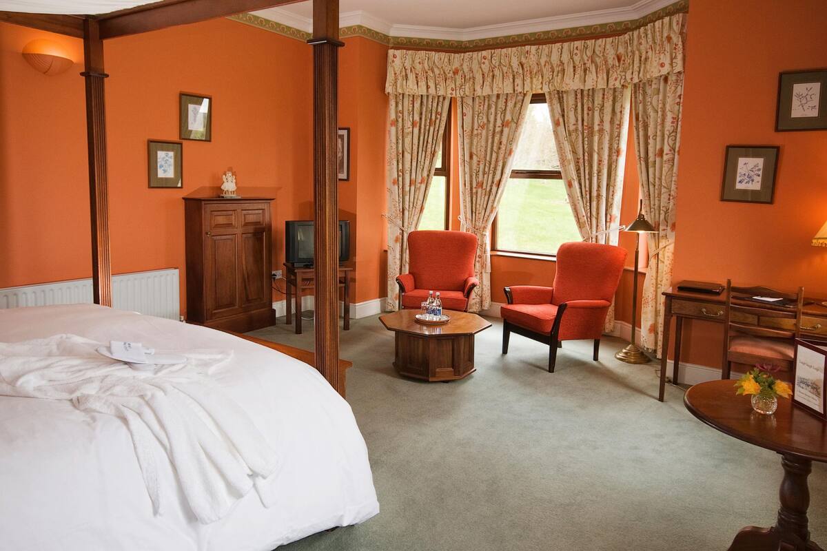A hotel room with a large bed, two red armchairs, a wooden coffee table, a desk with a lamp, and a view of the outdoors through a window with curtains.