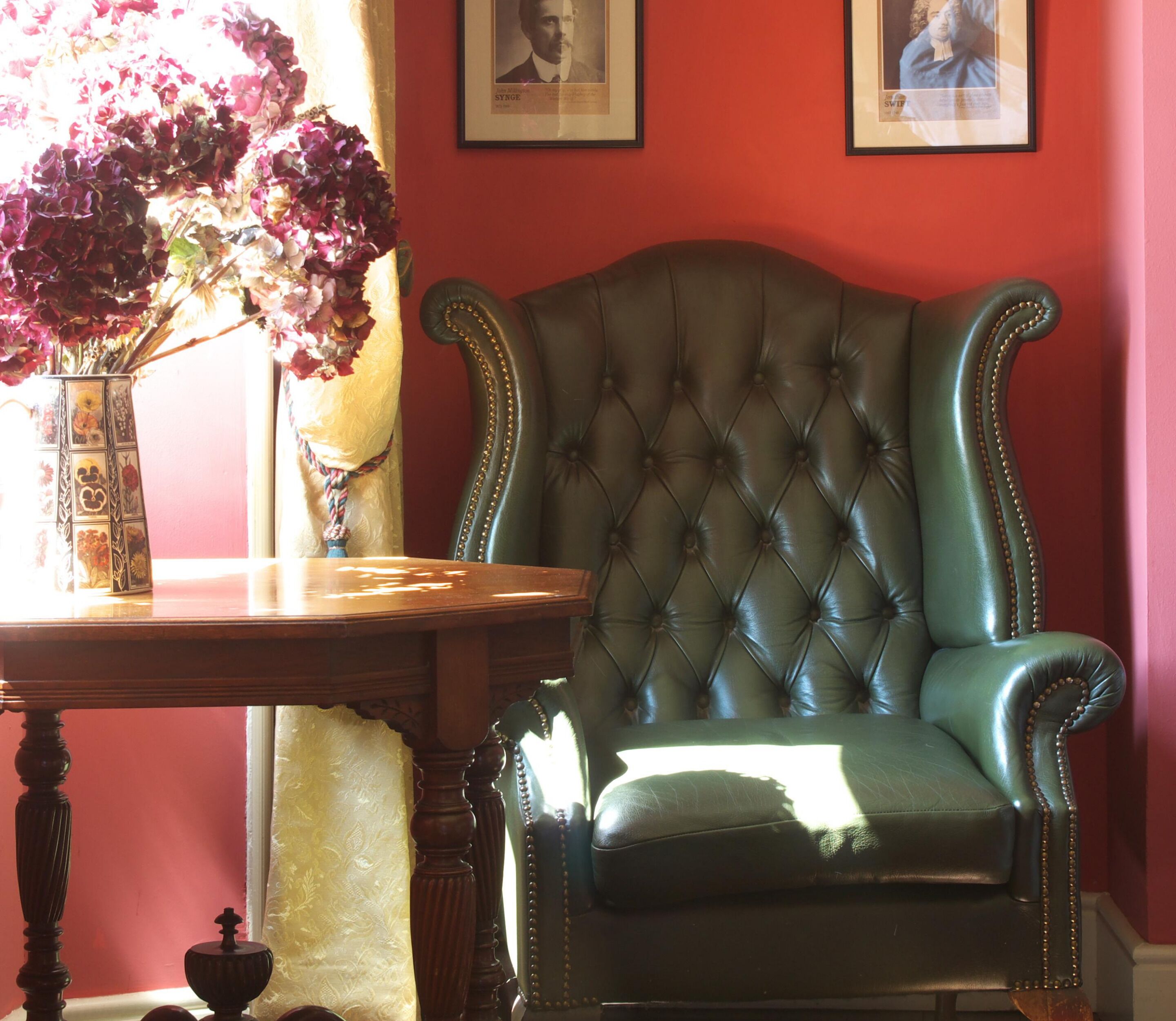 A room with a green upholstered armchair, a wooden table with a vase of flowers, and two framed pictures on the wall.