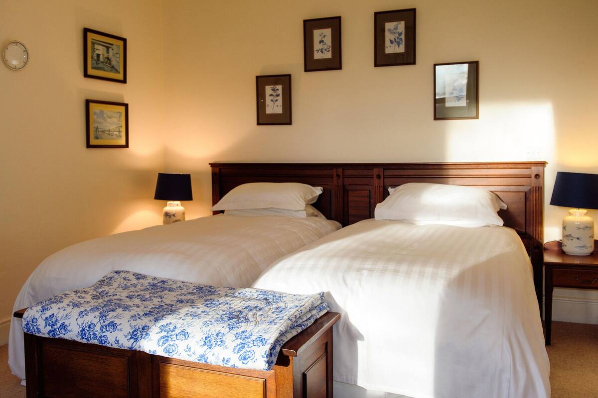 A double bed with white linens and a blue floral bedspread, flanked by two bedside tables with lamps, in a room with framed pictures on the wall.