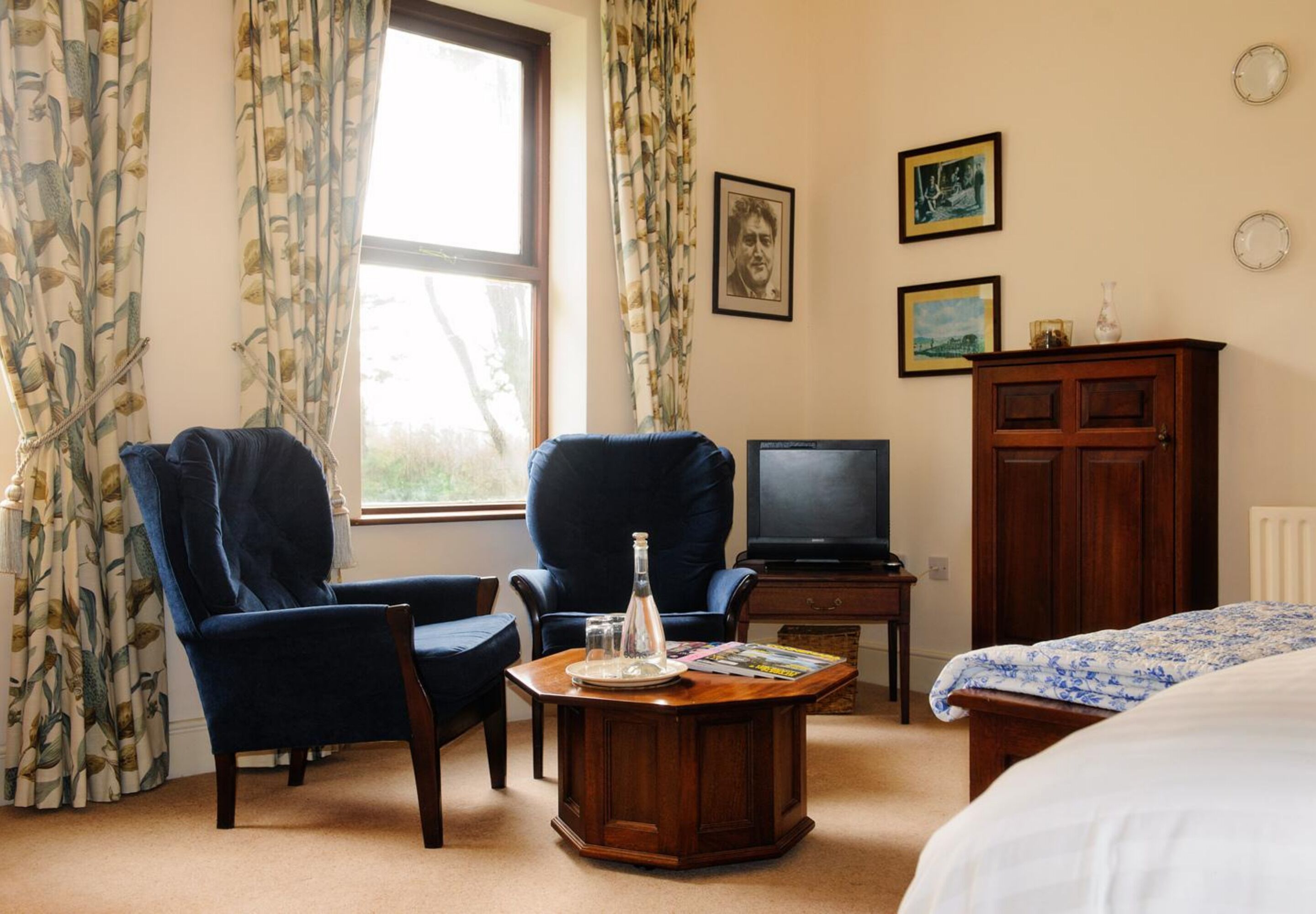 A hotel room with two armchairs, a wooden coffee table, a television, a cabinet, and a bed.