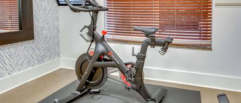 A stationary bike in a room with a patterned wall, a window with blinds, and a screen mounted on the wall.