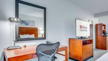 A hotel room with a wooden desk, a chair, a large mirror, a television, and a wardrobe.
