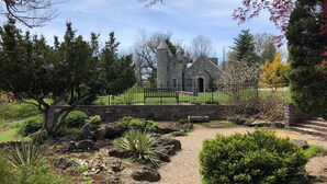 A stone building with a tower, surrounded by a well-maintained garden with various plants and a gravel path.
