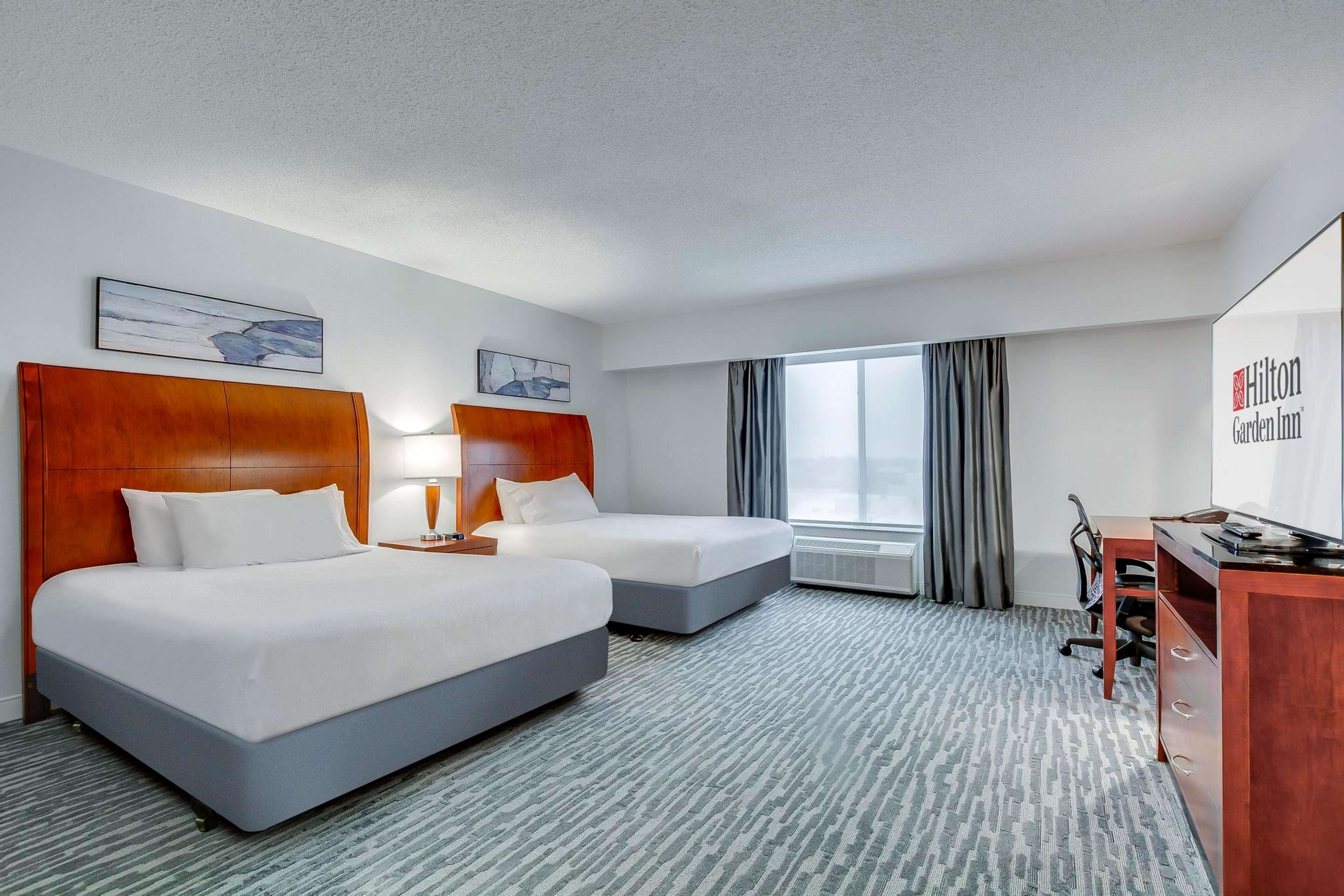 Quarto de hotel com duas camas, uma escrivaninha e uma janela grande com cortinas.