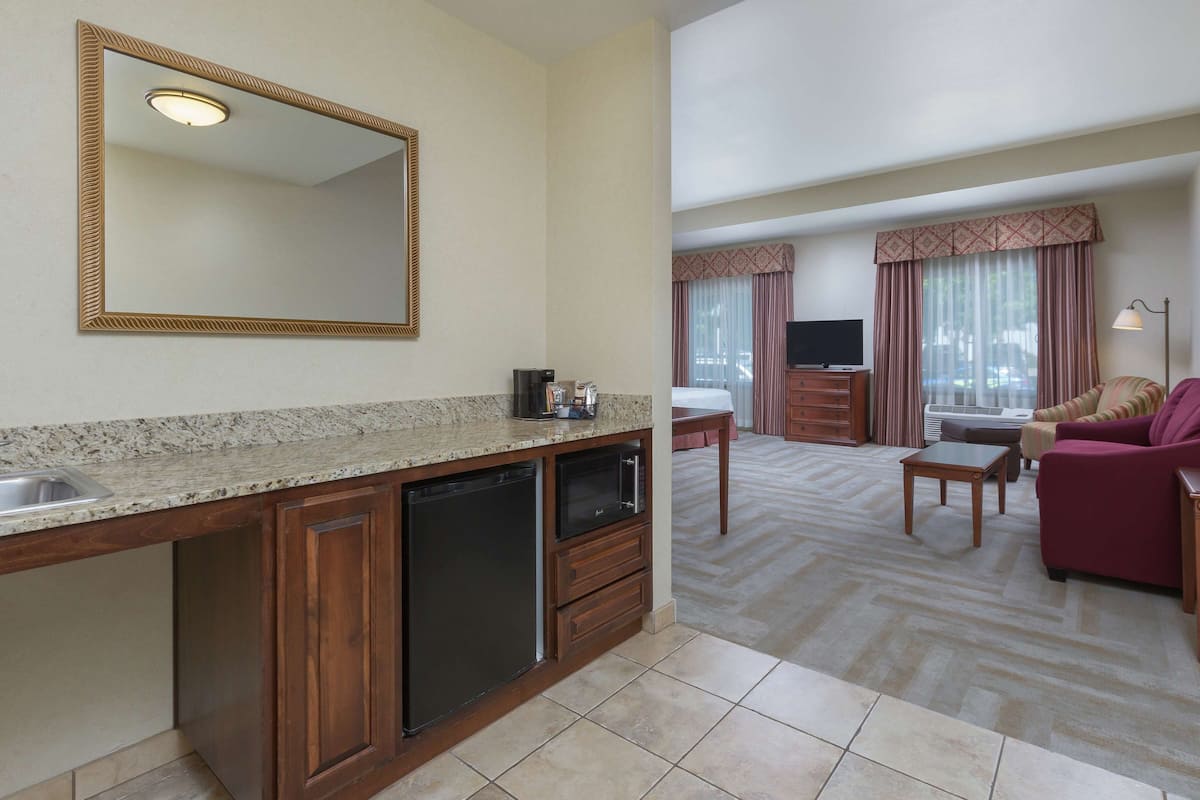 A hotel room with a kitchen area, a living room with a sofa and TV, and large windows with curtains.