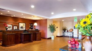 Zona de recepción de un hotel con un mostrador de madera, cuadros en la pared, una planta en maceta y un jarrón con girasoles.
