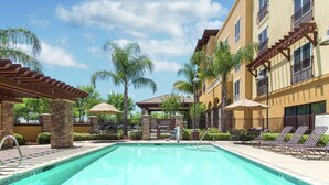 Una piscina con sillones de playa y sombrillas, rodeada de edificios con balcones y una pérgola.