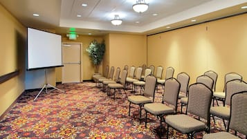 Un salón de conferencias con alfombra estampada, filas de sillas, una pantalla de proyector y una puerta con letrero de salida.