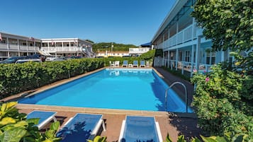 A swimming pool with lounge chairs, surrounded by greenery and a building with a balcony.
