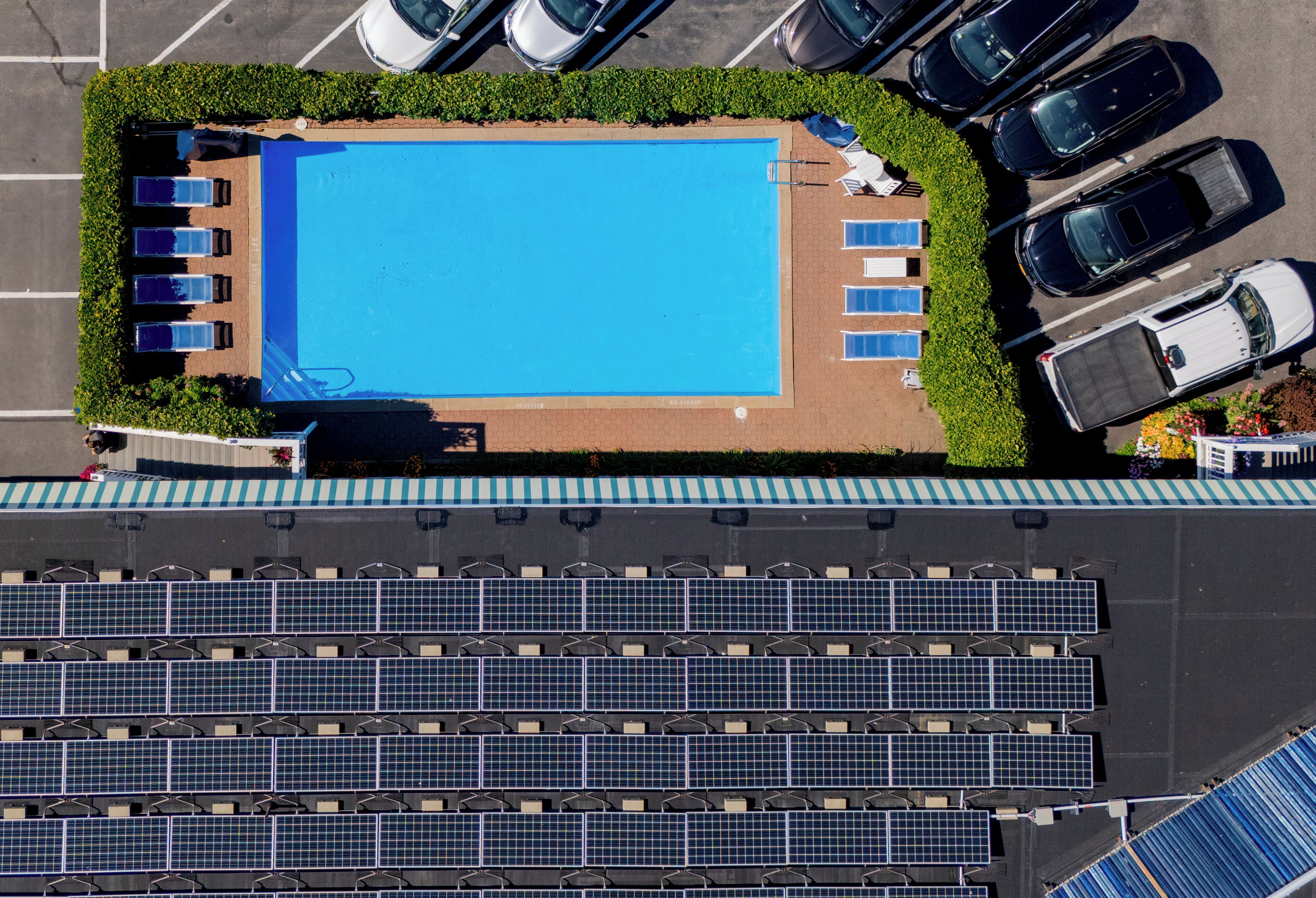 Vue aérienne d’un hôtel doté d’une piscine entourée de chaises longues et d’un parking où se trouvent des voitures et des panneaux solaires.