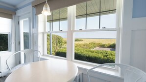 A room with a round table and white chairs, large windows with a view of a grassy field and trees, and a hanging light fixture.