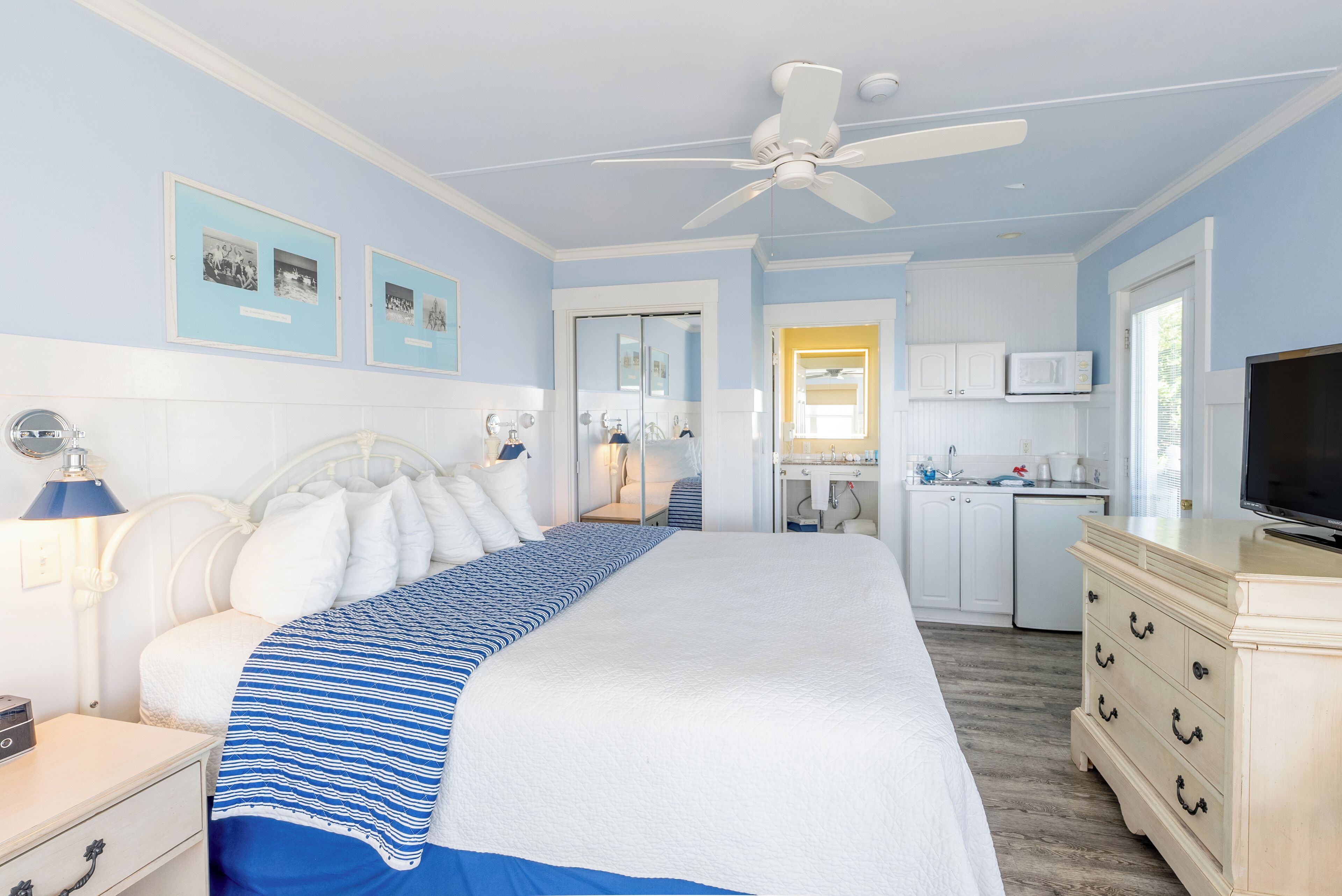 A bedroom with a large bed, a nightstand, a desk, a ceiling fan, a TV, and a kitchenette area.