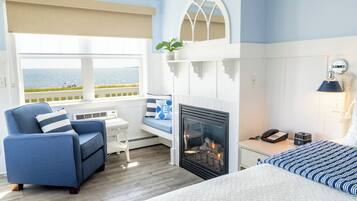Een hotelkamer met een groot bed, een blauwe fauteuil, een bijzettafeltje, een open haard, een nachtkastje en uitzicht op zee.