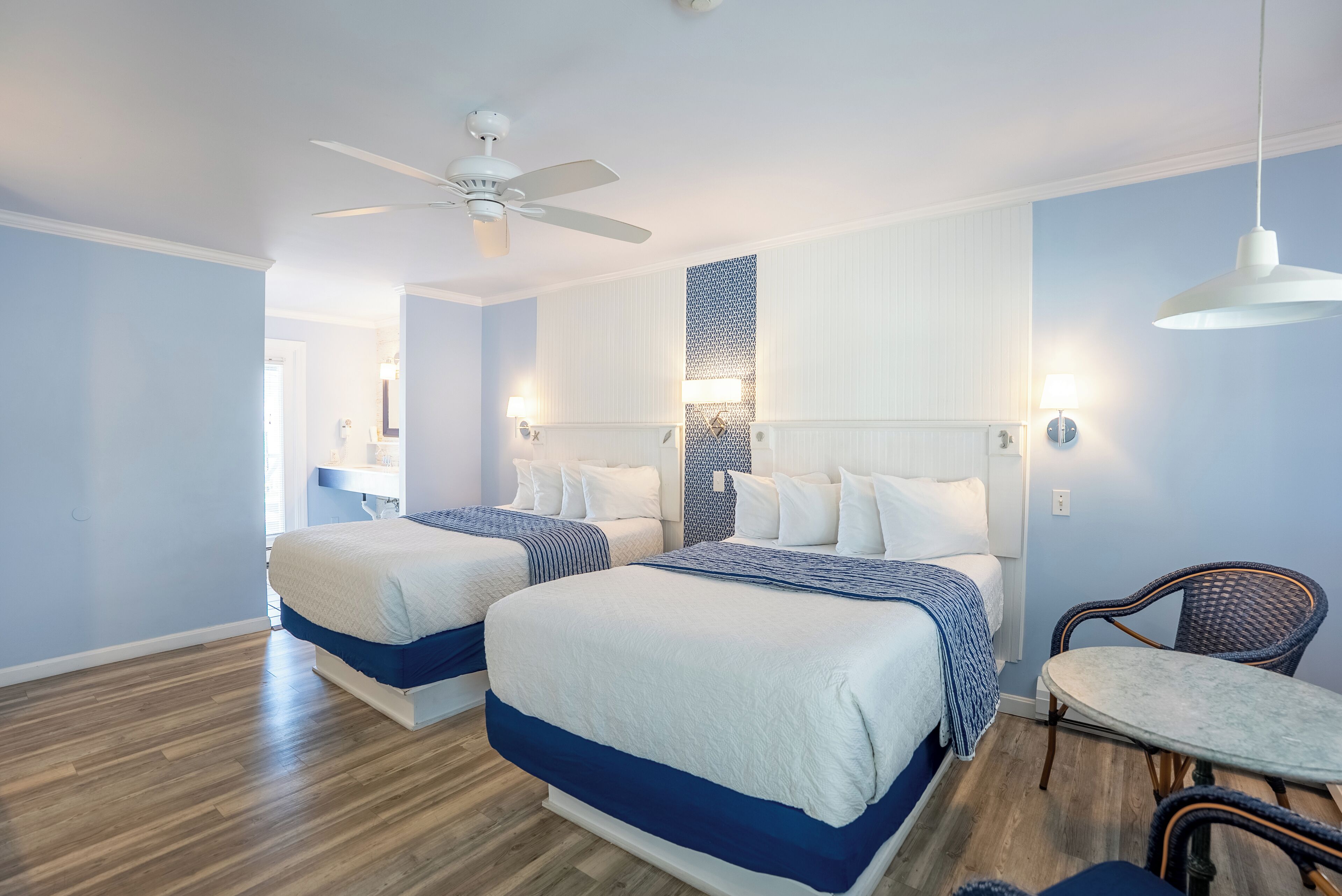 A hotel room with two beds, a ceiling fan, a table with chairs, and a wall-mounted light fixture.