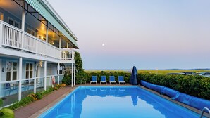 A pool area with a clear view of the ocean, a covered deck with lounge chairs, and a building with a balcony.