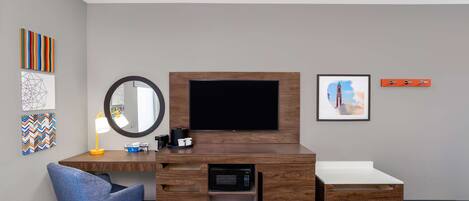 A hotel room with a wooden desk, a flat-screen TV, a mirror, a bed, and artwork on the walls.