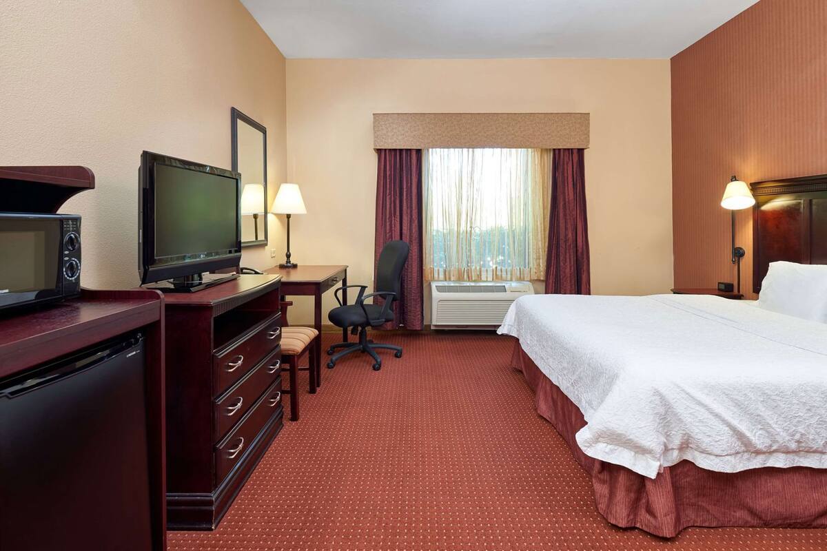A hotel room with a bed, a desk with a chair, a television, a dresser, and an air conditioning unit.