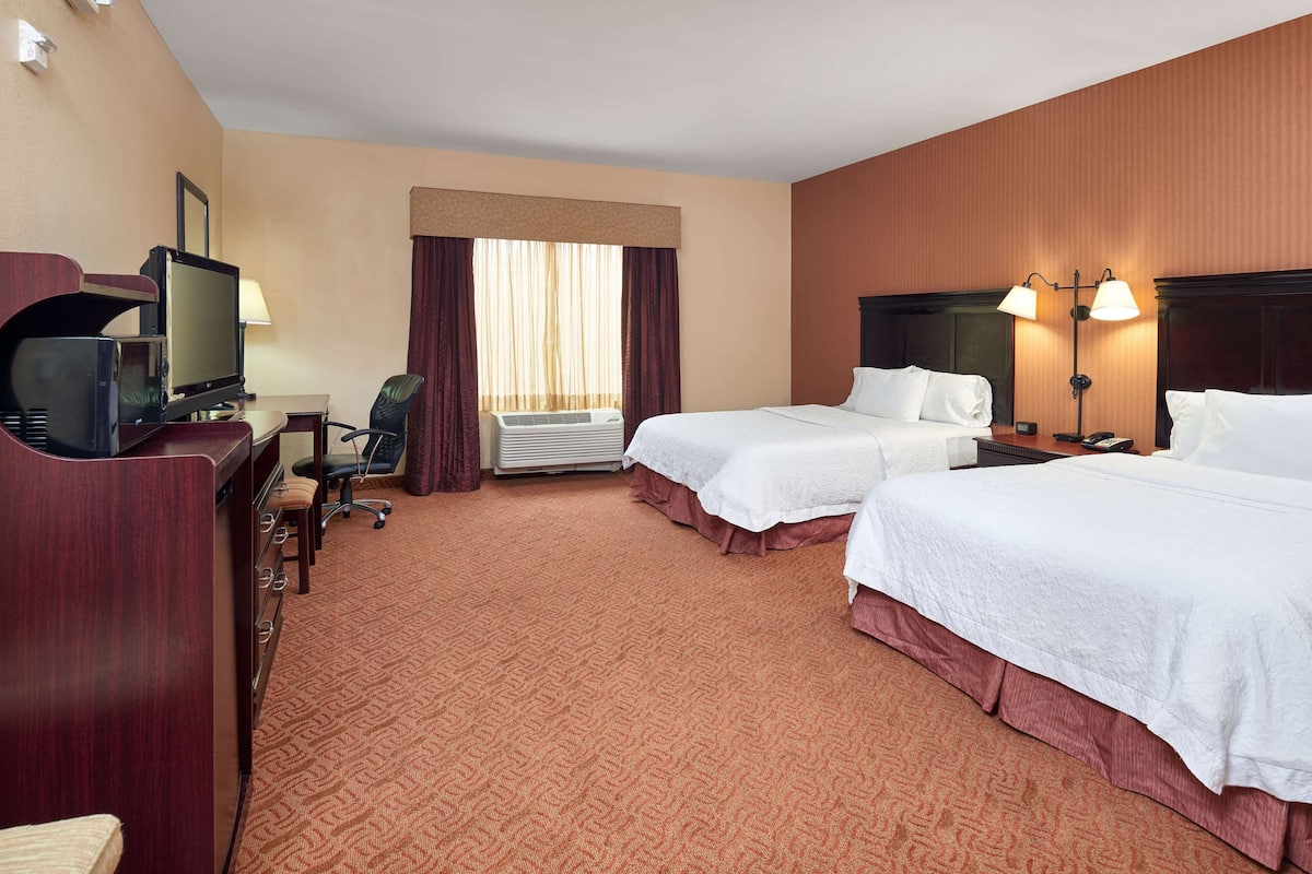 A hotel room with two beds, a desk with a television, a chair, and an air conditioning unit.