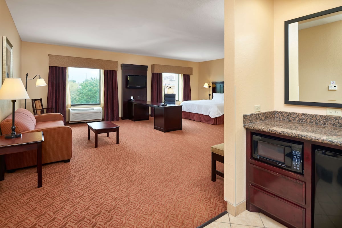A hotel room with a bed, a desk, a chair, a sofa, a TV, and a kitchenette.
