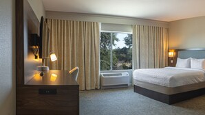 A hotel room with a bed, bedside tables, a desk, a chair, and a view of trees through the window. - Hampton Inn & Suites Boerne (Boerne)